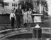 Drängen på Jonsjö säteri, August Gabrielsson, står på kanten till en gjuten damm omgiven av ungdomarna Jenny och Anna Lidberg samt Evert Klasson. I förgrunden en stor urna på sockel i dammens mitt och i bakgrunden säteriets huvudbyggnad.
(Se även bild EA2018)