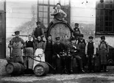 Ett antal anställda vid Alingsås bryggeri står uppställda vid tunnor och pumpar. I mitten står ett litet bord uppdukat med flaskor och glas. Vid bordet syns Theodor Möller till höger och Hjalmar Möller till vänster.