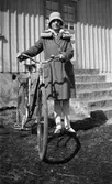 En flicka står med en damcykel vid stentrappan till ett hus. Hon är klädd i klockad hatt och kappa med pälskrage, vita strumpor och remskor.