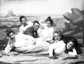 Ateljéfoto med en scen som ska ge sken av några vänner på stranden. Längst fram ligger Mathilda Ranch med en bok; bakom henne kollegan Elof Ernwald och kvinnan som ligger åt höger är fröken Augusta Pehrson (drev damskrädderi). Hon håller i en kartong märkt 