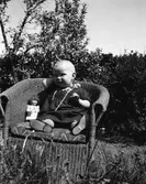 Ett litet barn med långt halsband sitter utomhus i en korgstol med en docka.
(Se även bildnr EA0891)