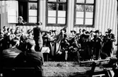 Orkester med sångare på friluftsmöte med Betaniaförsamlingen i Veddige. Bland instrumenten märks luta, banjo, mandolin, gitarr, fiol och dragspel. några barn lyssnar från ett öppet fönster i Betaniakapellet bakom orkestern.
(Se även bildnr EA0976 och EA2143)
