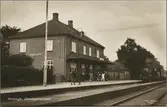 Spårsida av station med trafikerande persontåg och resande vid perrong.