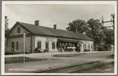Stationsområde med stationshus och elektrifierad bangård med resande på perrong.