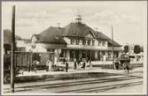 Vy på stationsområde med stationshus och resande i bild. Godsvagnen har littera Nässjö - Oskarshamns Järnväg, NOJ G 481.