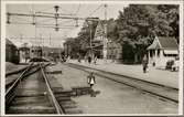 Vy på bangård och stationsområde med resande och lok i bild.