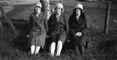 Tre unga kvinnor sitter på en grönskande dikesren, klädda i vårhattar, kappor, och strumpor.