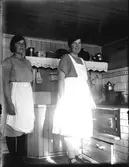 Två kvinnor vid järnspisen i ett lantkök. De har vita förkläden så kanske serverar de vid ett kalas. Runt spisen är det kaklat, väggen är spontad och vägghyllan är försedd med en broderad bård. På väggen hänger ett emaljkärl för såpa och ett av porslin för salt.