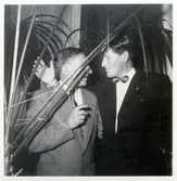 Två män står och pratar vid en fest anordnad av Monarks cykelverkstad för personalen. Ena mannen skyms av en större krukväxt. 
Lennart Eriksson, Idrottsmagasinet, till höger