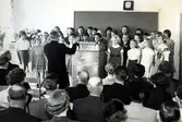 Brattåsskolans invigning 1954 eller 1955.
Folkskollärare och kantor Gunnar Alander leder en kör 
av elever. Föräldrar och politiker bland publiken.