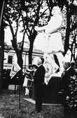 Riksdagens 500-årsjubileum firas. Carl Eldhs staty av Engelbrekt har avtäckts utanför Heliga Trefaldighetskyrkan. August Sävström, talman i riksdagens andra kammare, hyllar Engelbrekt.
Medeltidsklädda män med fanor skymtar i bakgrunden.