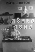 Karin Jonssons monter med läderarbeten. Här visas väskor, plånböcker och knappar av läder.
Karin bär ett långt halsband. På hennes monter står en vas med blommor.
(Karin är dotter till August Jonsson, som ägt skoaffären Jonsson & Blom och Gracilsko).