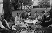 Avkoppling i Ahllöfsparken/Olof Ahllöfs park nära Arbogautställningen. De har kafferast i förberedelsearbetet inför utställningen.
Från vänster: Sven Drakenberg (intendent), Britta Orén, Olga Anderzon (från hemslöjdsföreningen), okänd, längst bak, i hatt,  Hanna Hartelius, Ulla Hermansson, Augusta Eriksson och Elin Höglund (konstnärinna)
Sju kvinnor och en man sitter på utlagda trasmattor. De har kaffekoppar och kaffekanna med sig.
