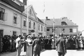 Människor är samlade på Stora torget i väntan på kung Gustaf V´s ankomst. Han, och en stor del av kungahuset, ska närvara vid Riksdagens 500-årsjubileum som firas i Arboga. Människor står bakom avspärrningar. Fönstren är öppna på pensionatet i Stadsgården så människor kan titta ut. I fastigheten på Västerlånggatan 1 sitter det till och med folk på taket. Flaggorna är hissade.
(Arbogautställningen pågår samtidigt).
