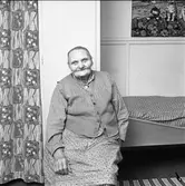 Kvinna, 90 år, iklädd väst och kjol. Hon sitter intill sin säng. På väggen, över sängen, hänger en bonad med katter. Till vänster, om kvinnan, hänger ett draperi.