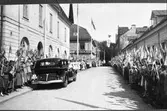 Arboga firar Sveriges Riksdags 500-årsjubileum. Kung Gustaf V, och en stor del av den kungliga familjen, är i staden med anledning av detta. Kungligheterna promenerar nu på Rådhusgatan medan skolbarn står på trottoarerna och vinkar med Svenska flaggan.
Konungens bil kommer att köra förbi Normans Efterträdare Damkonfektion, till vänster i bild.
