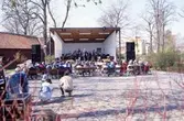En blåsorkester spelar på scenen i Olof Ahllöfs park. Publiken sitter på bänkar och lyssnar.