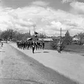 Arboga Blåsorkester marscherar på Kungsörsvägen. I täten går två män med var sin fana.
I bakgrunden syns tornet på Heliga Trefaldighetskyrkan och till höger om det reser sig skorstenen på Arboga kvarn. Till höger, i bild, skymtar hamnen och järnvägen.