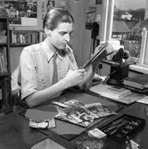 Arboga Tidning, interiör och personal.
En man sitter vid ett skrivbord. Han är iklädd skjorta och slips. Han har en cigarett i munnen. På skrivbordet finns ett cigarettpaket och ett askfat. Mannen klipper med en lång sax. Framför honom, på skrivbordet, ligger bilder i olika format. På skrivbordet står en telefon. På väggen hänger en annan telefon, kanske en interntelefon? På väggen, bakom honom, finns en mängd bilder.  Skrivbordet står intill ett fönster. Mannens kontor ligger någon våning upp i huset.