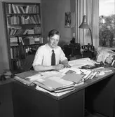 Arboga Tidning, personal och interiör.
En man sitter vid ett skrivbord med en penna i handen. Möjligen är han journalist. Han har skjorta och slips på sig, kavajen har han hängt över stolsryggen. Skrivbordet är belamrat med papper och trycksaker. Där finns även en telefon, en askkopp och en skrivbordslampa. Skrivbordet står nära fönstret, det syns att mannens kontor ligger någon våning upp i huset.
Vid sidan om honom, står ett litet bord med en skrivmaskin på. På väggen hänger en telefon (möjligen en interntelefon). Bakom sig har han en bokhylla full av böcker och pärmar. Det står till och med några böcker på golvet.