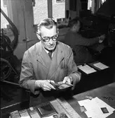 Arboga Tidning, personal och interiör.
Två män, båda i skyddsrock, arbetar på var sin arbetsplats. Mannen närmast kameran är möjligen sättare. Han har en sax i handen. På bordet, framför honom, ligger några porträttbilder. Mannen i bakgrunden arbetar vid en maskin.