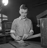 Arboga Tidning, personal och interiör.
En ung man arbetar vid ett bord. Möjligen är han typograf? Han har ett pincettliknande verktyg i handen.