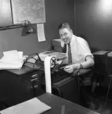 Arboga Tidning, personal och interiör. En man, iklädd skjorta och slips, arbetar vid en räknemaskin. Kontorsstolen är försedd med hjul. På skrivbordet ligger en uppslagen pärm. På väggen hänger en karta.