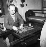 Arboga Tidning, personal och interiör. En man, iklädd skyddsrock, sätter en tidningssida. Han har trycksvärta på fingrarna. På väggen bakom honom finns en strömbrytare.