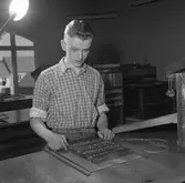 Arboga Tidning, personal och interiör.
En ung man arbetar i tryckeriet. Han bygger en tidningssida. På bordet, framför honom, ligger typer i olika storlekar. Bakom ryggen har han ett fönster. Det syns att han arbetar på andra våningen i huset.
Fotografens assistent håller i blixten och dennes hand syns i bildens övre, vänstra hörn.