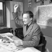 Arboga Tidning, personal och interiör. En man, iklädd kavaj, sitter vid ett skrivbord och gör en segergest. Han håller ett papper i handen. På bordet ligger en tidning, ett kuvert och en stor sax. Vid sidan står en skrivmaskin på ett lågt bord. På väggen, bakom mannen, hänger en karta och små fotografier.