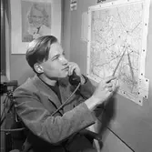 Arboga Tidning, personal och interiör. En ung man, iklädd kavaj, sitter på kontoret. Han har en telefonlur i ena handen och en pekpinne i den andra. Han pekar på en karta som är uppsatt på väggen. På väggen, bakom hans rygg, sitter en bild på en ung flicka.