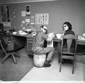 Arboga Tidning, personal och interiör. Två journalister arbetar vid samma skrivbord.  De vänder sig om, mot fotografen. Mannen sitter på en papperskorg och kvinnan på en trästol. Till vänster om dem finns en obemannad arbetsplats med en telefon och en arbetsstol på hjul. En skrivmaskin skymtar. På väggen sitter bland annat en affisch med olika svampar.