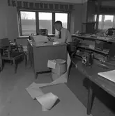 Arboga Tidning, personal och interiör.
En man arbetar vid sitt skrivbord. Han har en flera meter lång utskrift, delvis liggande på golvet. I förgrunden står en kamera. Mannen sitter på en kontorsstol på hjul. På andra sidan skrivbordet står en karmstol till besökare. Bakom stolen står en skrivmaskin på ett lågt bord. Skrivbordet står intill fönstret, det syns att kontoret ligger på andra våningen. Bakom mannens rygg finns en låg hylla som är full av papper och tidningar.