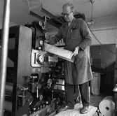 Arboga Tidning, personal och interiör. En man, iklädd skyddsrock, arbetar vid tryckpressen. Han håller en tidningssida med typer, i händerna. I fickan, på rocken, har han två pennor. På golvet, nedanför maskinen, ligger en rulle tidningspapper med tryck på. I bakgrunden skymtar en sopsäck.