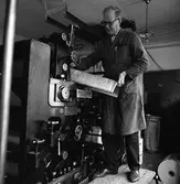 Arboga Tidning, personal och interiör. En man, iklädd skyddsrock, arbetar vid tryckpressen. Han håller en tidningssida med typer, i händerna. I fickan, på rocken, har han två pennor. På golvet, nedanför maskinen, ligger en rulle tidningspapper med tryck på. I bakgrunden skymtar en sopsäck.