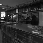 Arboga Tidning, personal och interiör. Flera personer ses arbeta i tryckeriet. Tryckpressen i bakgrunden. I förgrunden står en arbetsbänk med typer i lådorna.