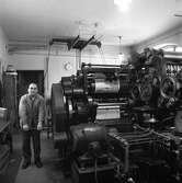 Arboga Tidning, personal och interiör. En man håller i en vev till en stor tryckpress. De färdigtryckta tidningssidorna syns på valsarna.