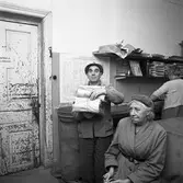 Arboga Tidning distribueras. En man håller en trave tidningar i famnen. I bakgrunden syns en kvinna som packar tidningar. I förgrunden sitter en kvinna, klädd i ytterkäder. Möjligen väntar hon på tåget. På väggen finns TGOJ´s tågtidtabell. Dörren, till vänster, är mycket sliten.