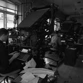 Arboga Tidning, personal och interiör. Två män sitter vid var sin sättningsmaskin. Maskinerna har tangentbord som männen skriver på. Handskrivna lappar ligger vid deras sida.