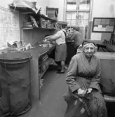 Arboga Tidning ska distribueras. Två kvinnor hanterar tidningsrullar vid en arbetsbänk. Mannen i hatt har anknytning till Arboga Tidning. På väggen hänger TGOJ´s turlistor. Är bilden tagen på järnvägsstationen? Kvinnan i förgrunden ser ut att vara på väg till arbetet. Hon har en matsäcksväska i handen. Hon är klädd i mössa och en kappa med svångrem i midjan.