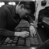 Arboga Tidning, personal och interiör. En man arbetar med typer vid en maskin. Han har en pincett i handen. På väggen ses en sax och en strömbrytare. Lokalens höga fönster syns i bakgrunden.