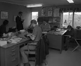 Arboga Tidning, personal och interiör. Fem personer på kontoret. Tre journalister arbetar vid sina skrivmaskiner. En person sitter vid skrivbordet och läser anteckningar. En man står upp och har ett långt papper i handen. På skrivborden syns telefoner, askfat och tidningar. På väggen, mellan fönstren, hänger bio- och teater affischer.