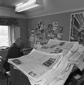 Arboga Tidning, personal och interiör. Journalisten vid sitt skrivbord på sportredaktionen. På väggen, framför skrivbordet, sitter diverse sportfotografier och andra bilder. I förgrunden ses två högar med tidningar.