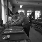 Arboga Tidning, personal och interiör. Två män som bygger tidningssidor med typer. Deras titel kan vara sättare. På väggen hänger en telefon. I förgrunden ses ett askfat. I bakgrunden står en stor maskin.