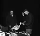 Arboganatta, orienteringstävling. Möjligtvis prisutdelning. Två män iklädda ytterkläder. I bakgrunden anas en bil.
På prisbordet finns bland annat en badrumsvåg, en uppsättning bestick, ett halvt dussin glas och en uppstoppad fågel.