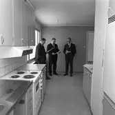 Arbogavillan lll, interiör, kök. Diskbänk, spis och köksfläkt i förgrunden. Tre kostymklädda herrar står i kökets matplats.
I mitten står Hilding Lund på Byggnadsfirma Lund.