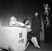 Medlemmarna i Arbogarevyn spelar upp sin Jubileumsrevy. Tre figurer på scenen, en ligger i vagga.