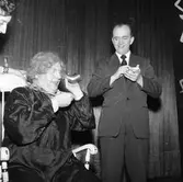 Arbogarevyns medlemmar spelar upp sin Jubileumsrevy. I en gungstol sitter en man utklädd till en äldre dam. Han använder hörlur. Mannen i kostym är Rune Ström.