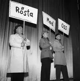 Arbogarevyns medlemmar sätter upp sin Jubileumsrevy. Tre män på scenen. De bär plakat och ytterkläder.