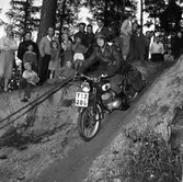 Arbogas Stjärnknutte
Tävling i terrängkörning med motorcykel. Man iklädd skinnluva och med vindglasögonen i pannan styr sin motorcykel i nerförsbacken. Motorcykeln har packväskor. Publiken står alldeles intill.